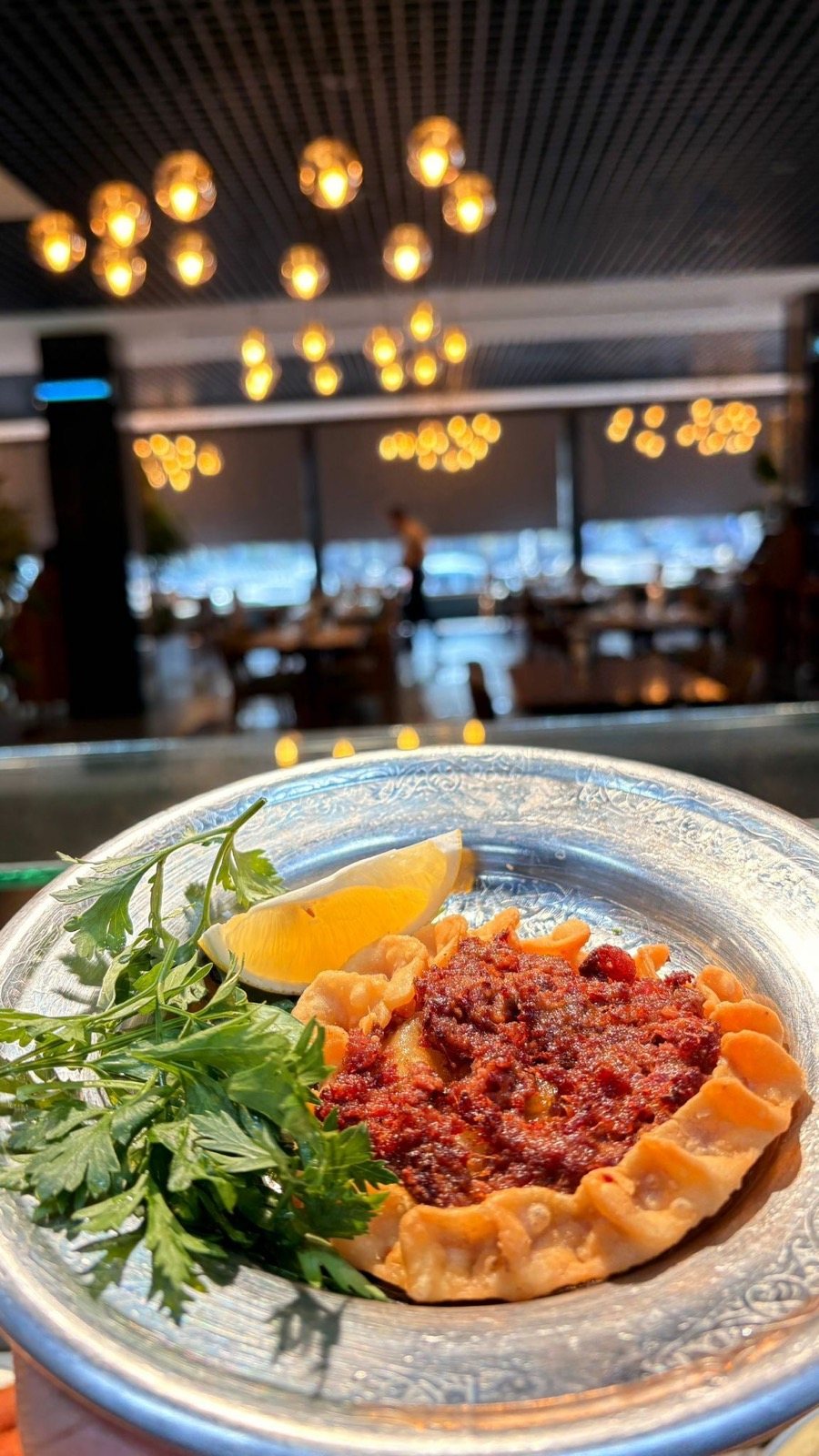 Gümüş tabakta ağzı açık içli köfte, limon ve maydanoz, arkada restoran salonu