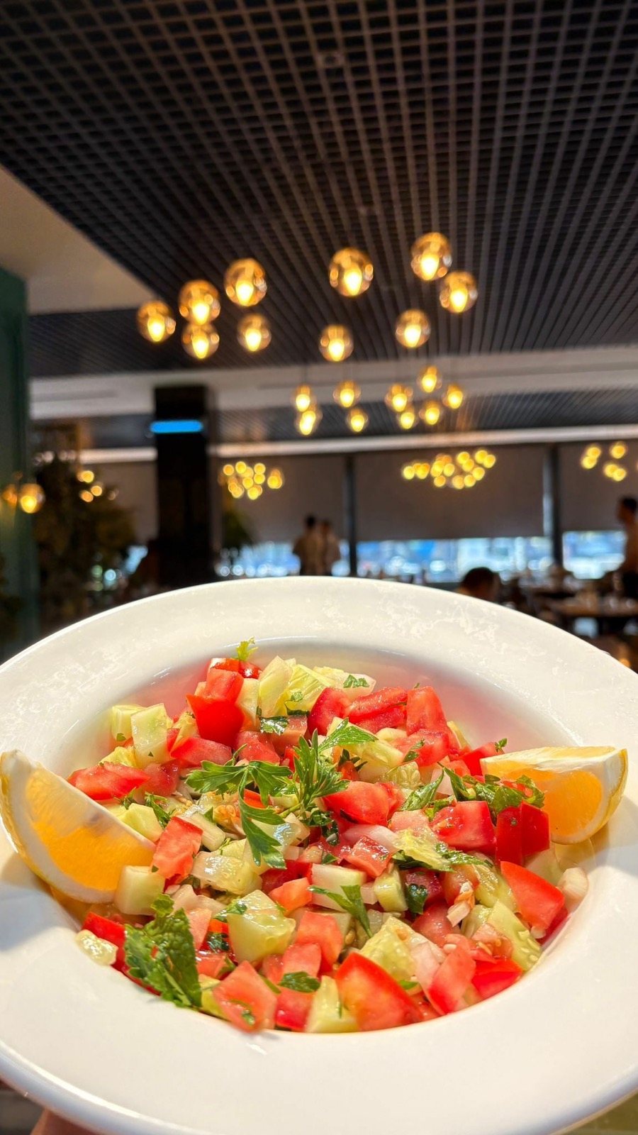 Beyaz kâsede çoban salatası ve limon dilimleri, arkada restoran salonu